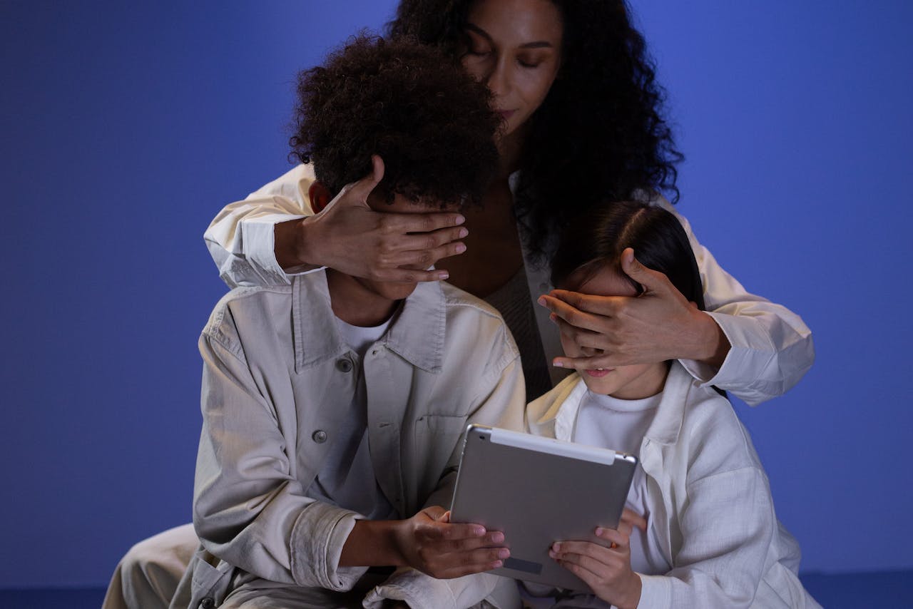 A woman shields the eyes of a boy and a girl looking at a tablet.