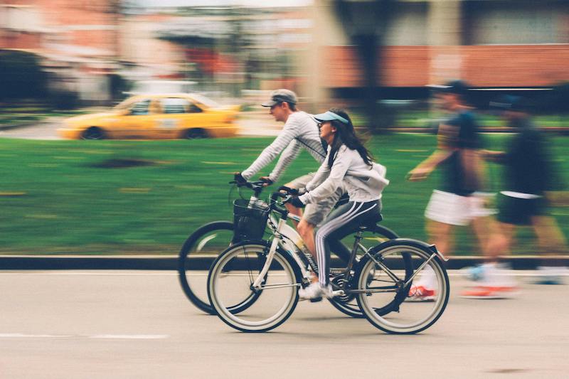 A man and a woman ride bikes with a pedestrian and cars in the background.