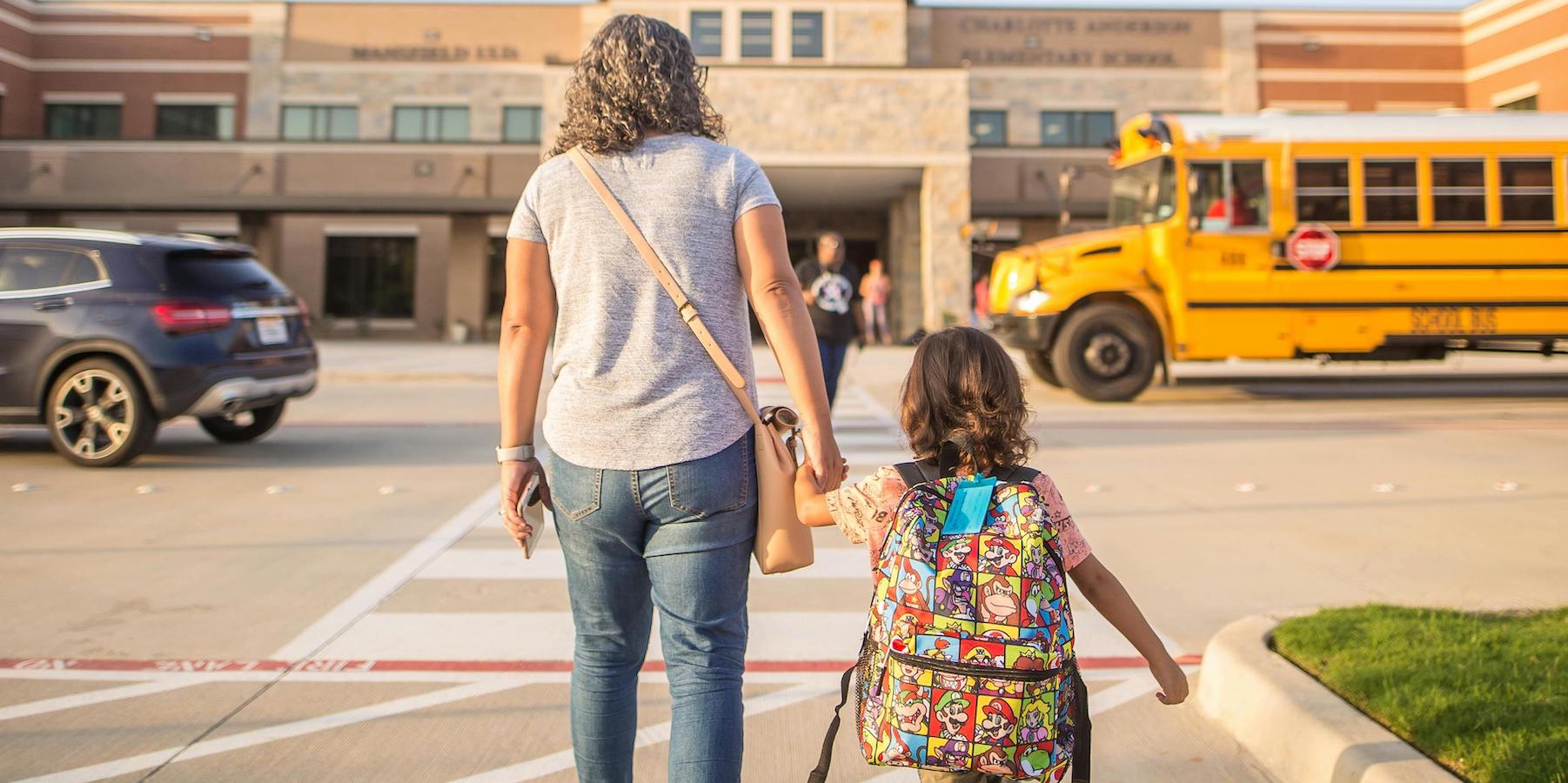 A woman walks a young girl across a crosswalk toward school, with a school bus in the background.