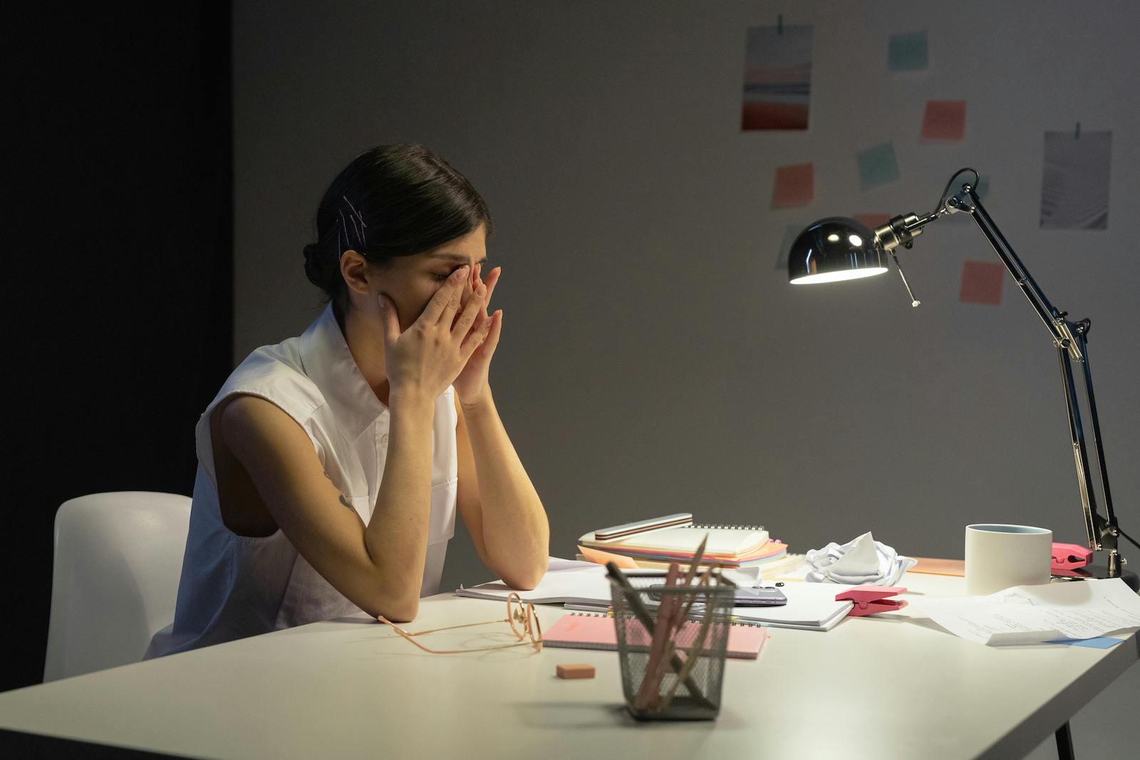 A woman with dark hair and a white blouse sits at a desk with her hands to her face.